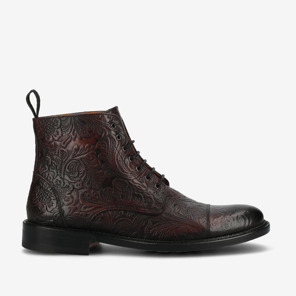 Side view of a dark brown, intricately embossed leather ankle boot with a black sole, lace-up front, and pull tab at the back.