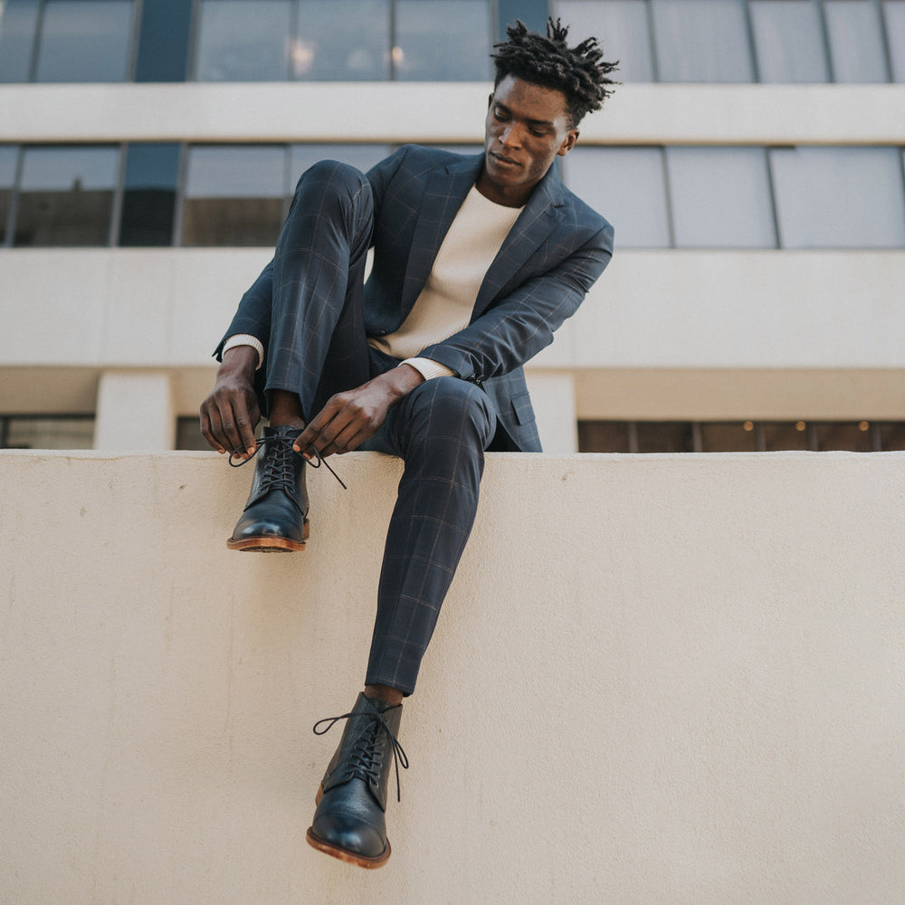 A man in a blue suit and white shirt sits on a ledge tying his black boots, with a modern building in the background.