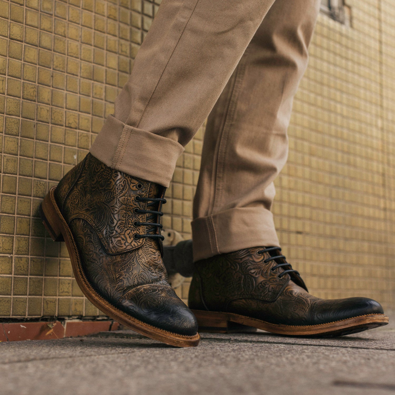 Person wearing tan pants and dark leather brogue boots with intricate patterns, standing on a tiled sidewalk.