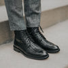 Person wearing woven black leather brogue boots and gray pants, standing on a concrete step.