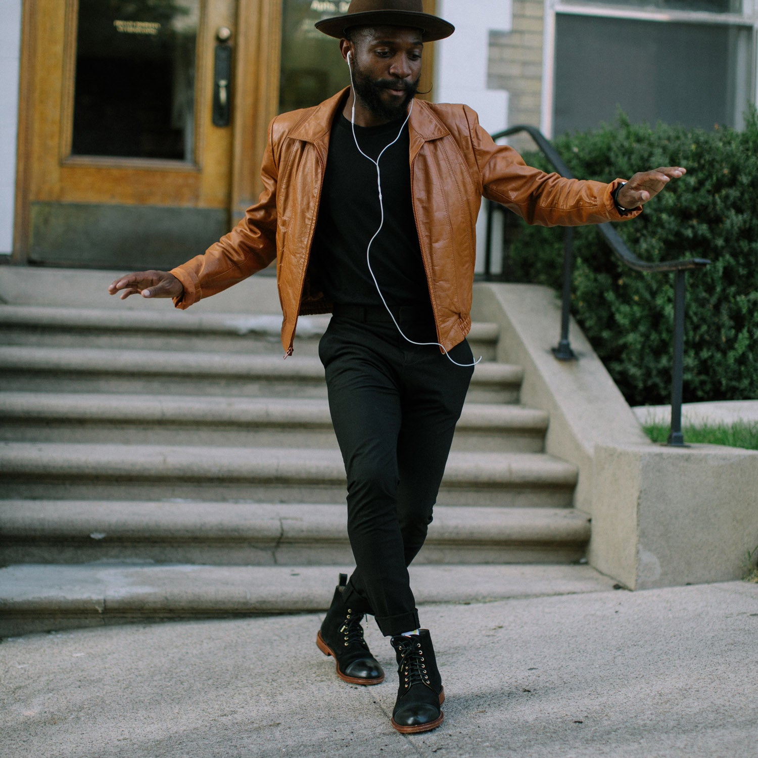 A man wearing a brown hat and leather jacket listens to music on earphones while dancing on a sidewalk near concrete steps.