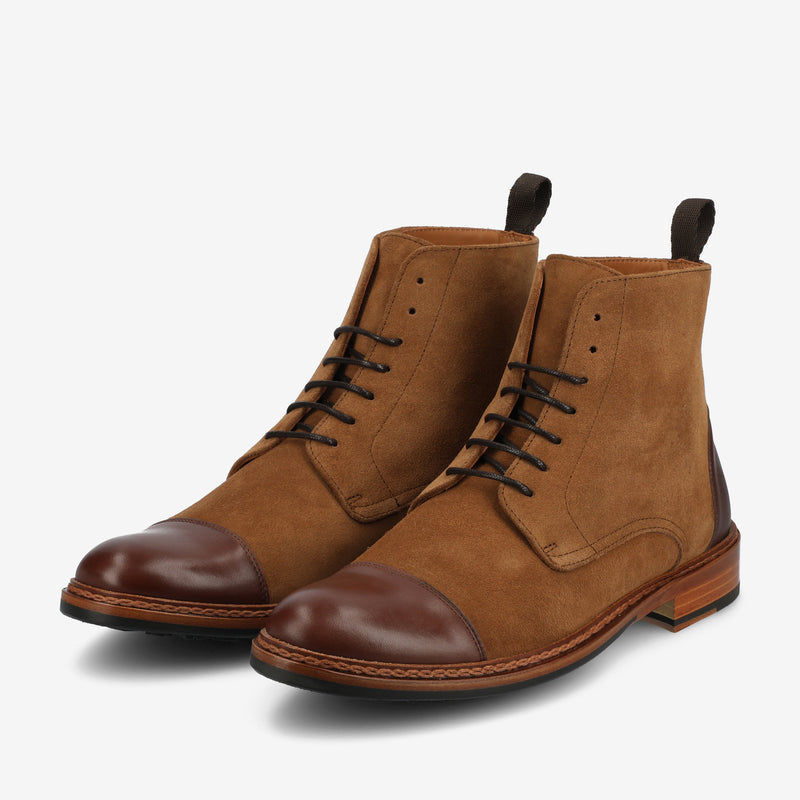 A pair of brown suede and leather cap-toe lace-up boots with dark brown soles and pull tabs at the back.