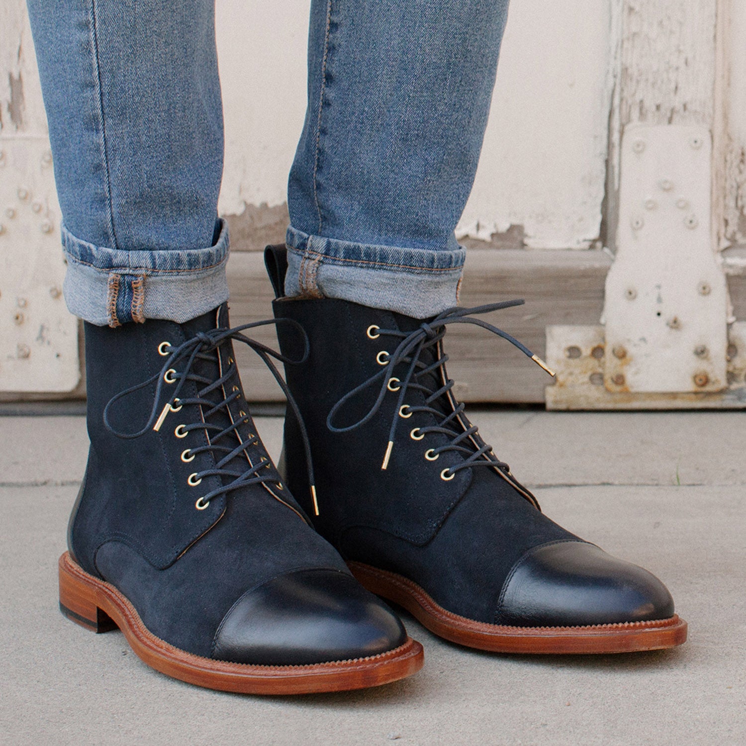 Person wearing rolled-up blue jeans and black leather lace-up boots with brown soles standing on a concrete surface.