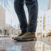 Close-up of a person wearing dark green leather boots with dark blue pants. The background shows an outdoor urban setting with buildings and cars.