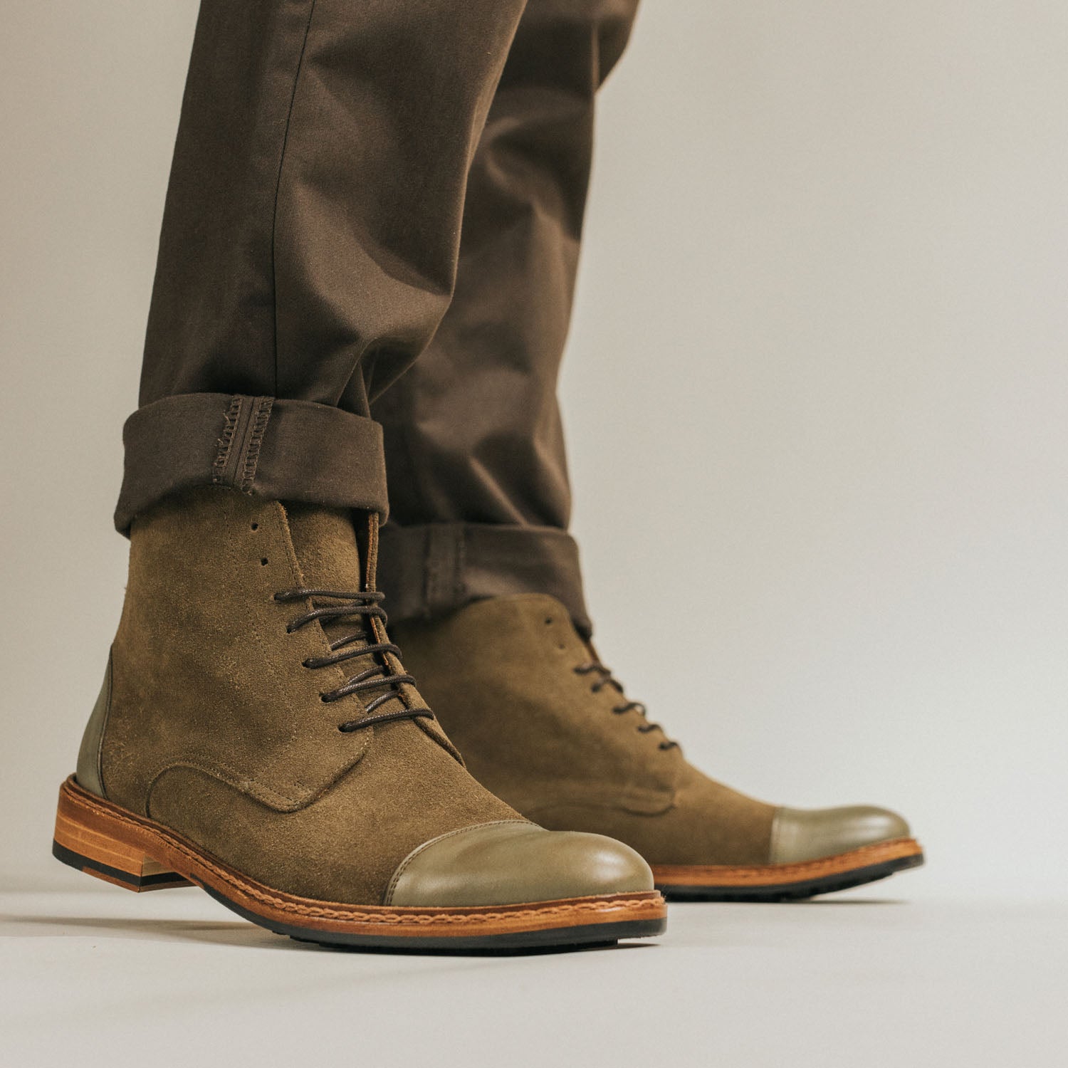 Close-up of a person wearing olive-green suede boots with light brown laces, paired with rolled-up brown pants. The shoes have a polished leather toe cap and a light wooden sole.