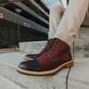 A person wearing burgundy and black leather boots sits on concrete steps, with beige pants slightly rolled up.