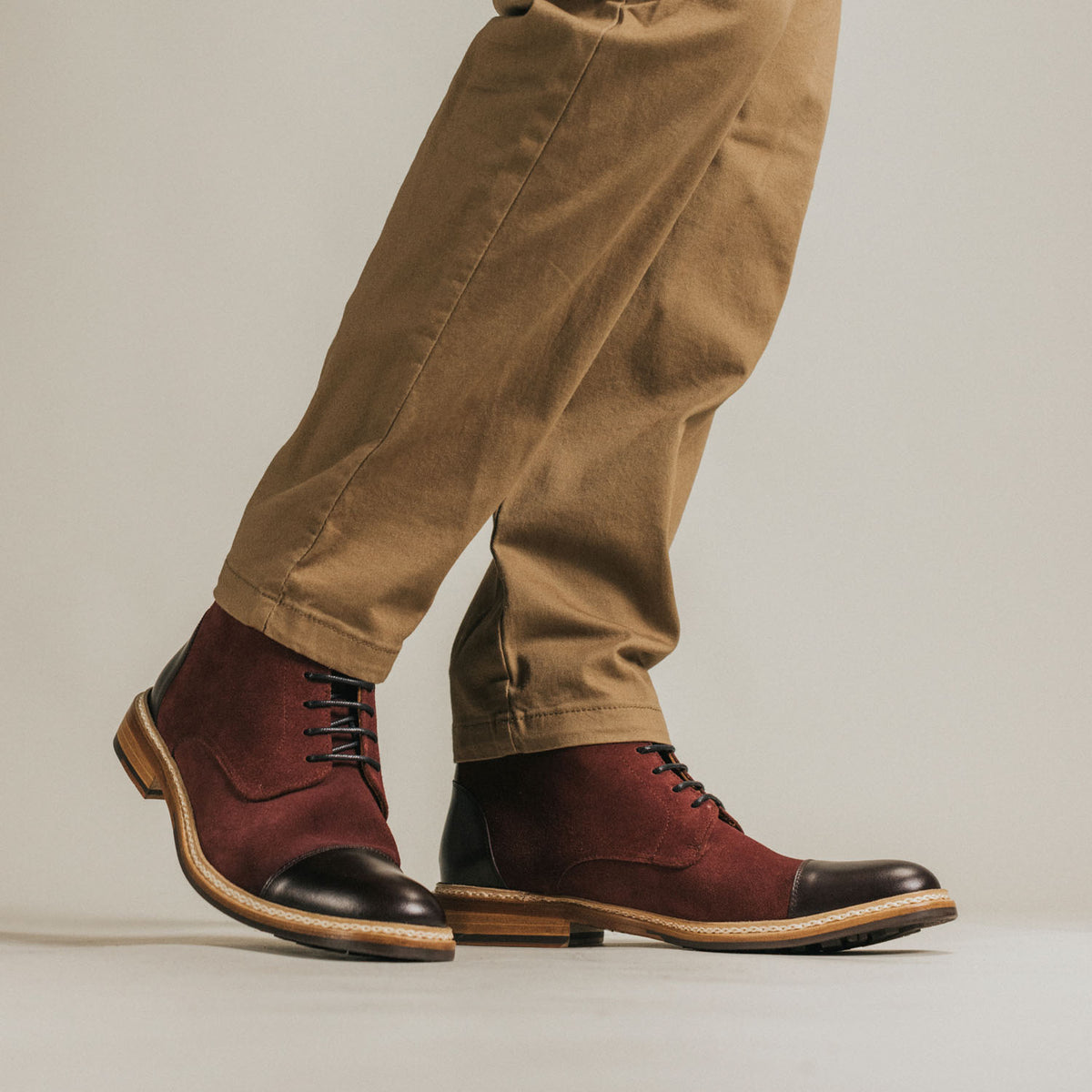 A person wearing brown pants and red-brown leather boots with black toe caps and heels stands on a light-colored floor.