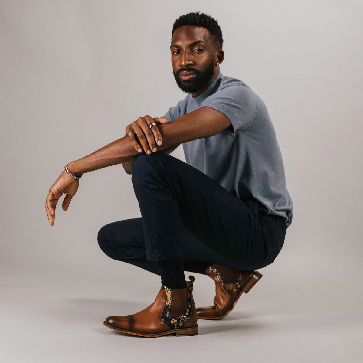 A man with a beard crouches on the floor, wearing a gray shirt, black pants, and brown leather boots.