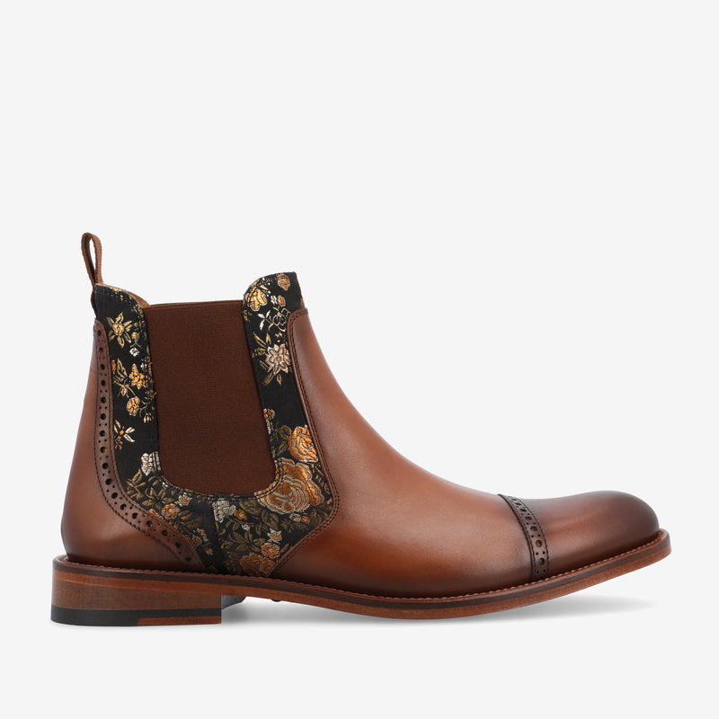 A brown leather Chelsea boot with patterned elastic side panels, featuring a pull tab and decorative stitching.