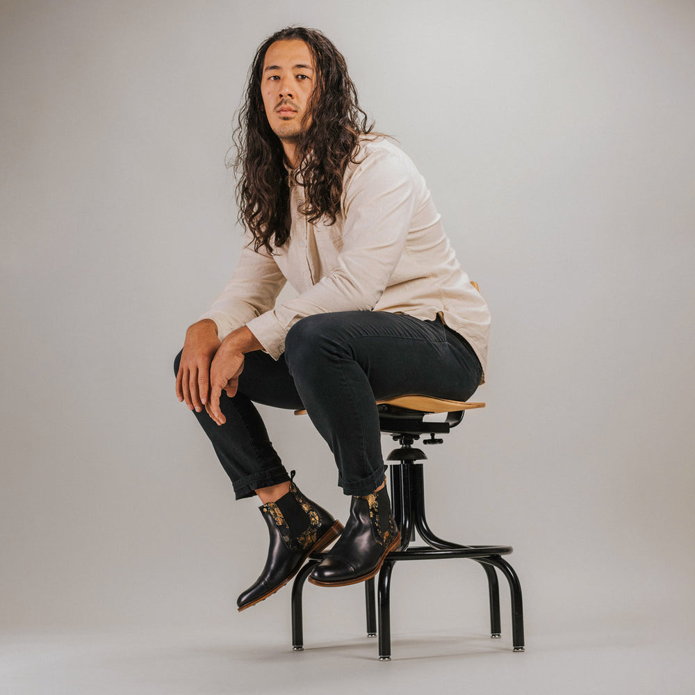 A person with long hair and casual attire sits on a stool. They are wearing a beige shirt, black pants, and black boots with a patterned design.