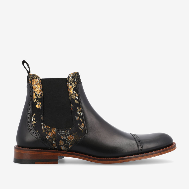 A black leather Chelsea boot with floral-patterned elastic side panels, a pull-tab at the back, and a brown wooden sole. The boot features decorative brogue detailing along the edges.