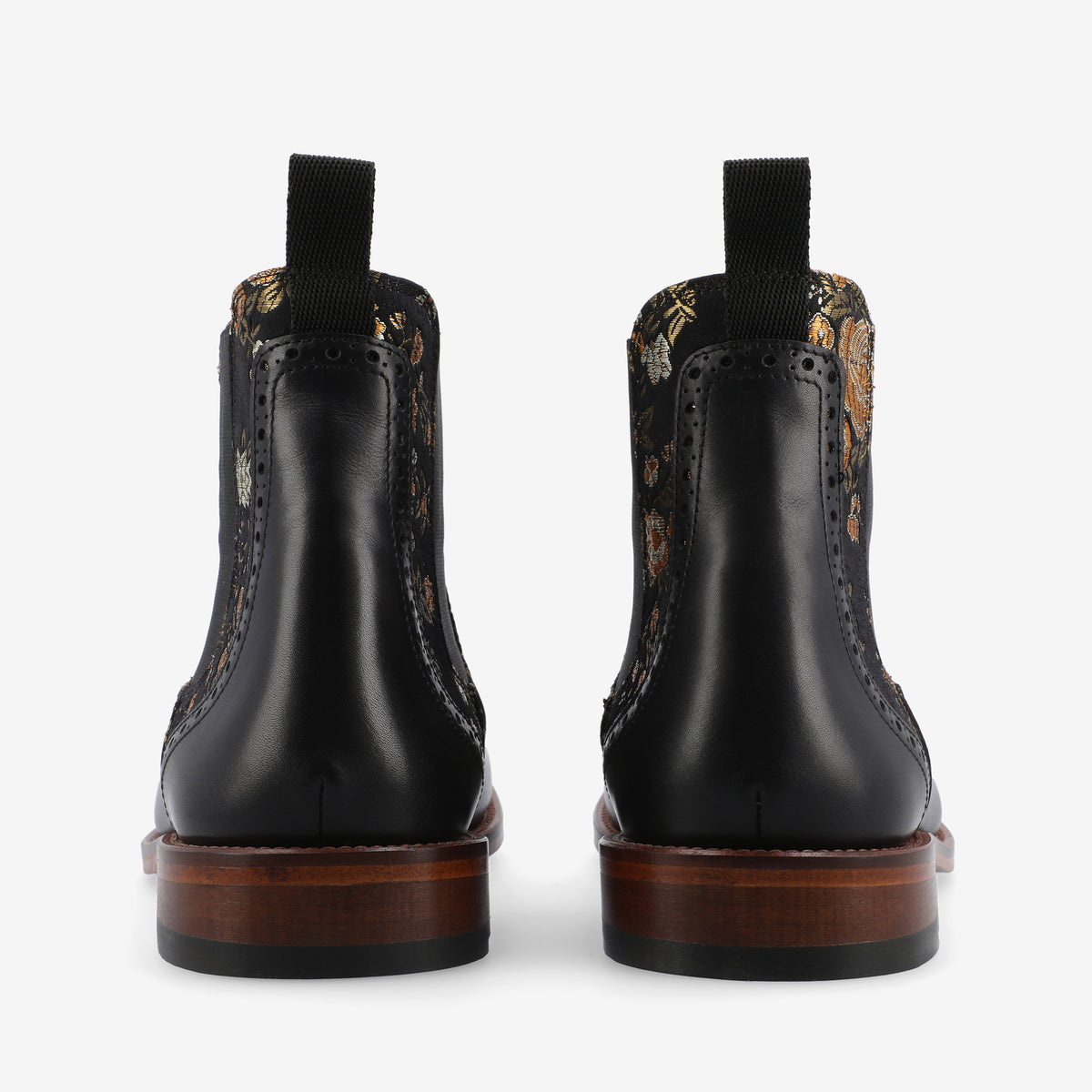 A pair of black ankle boots with ornate floral embroidery, black pull tabs, brown wooden soles, and brogue detailing viewed from the back.