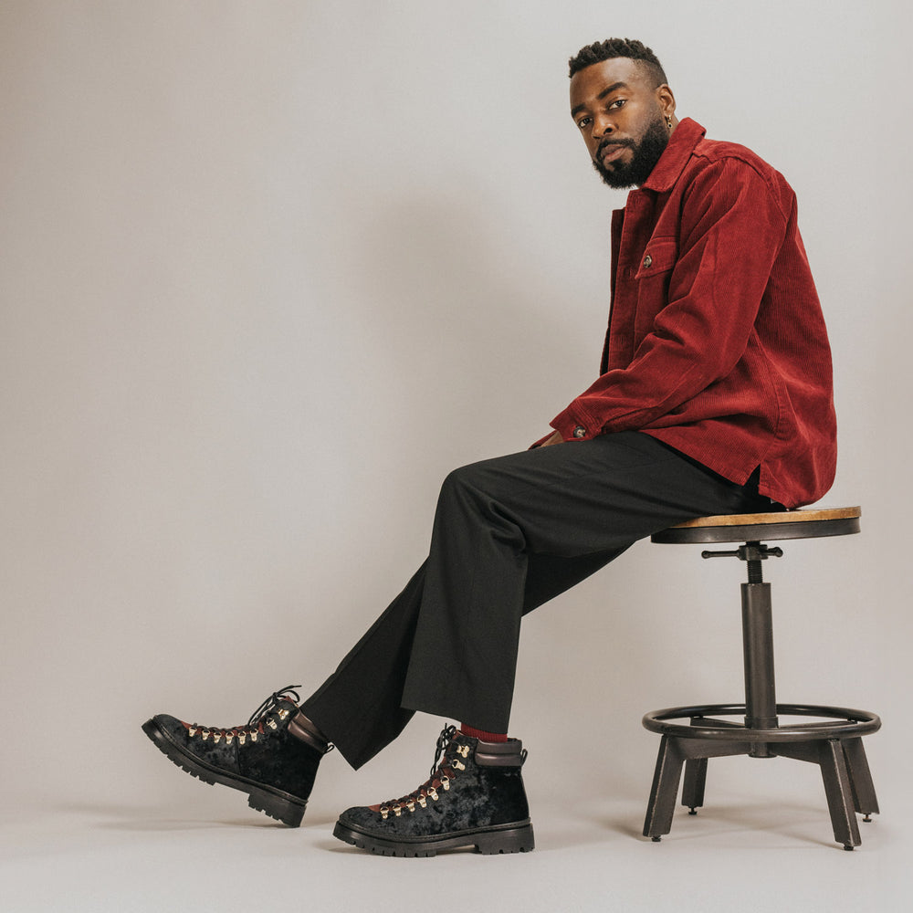 A bearded man wearing a red jacket, black pants, and black boots sits on a stool against a plain background.