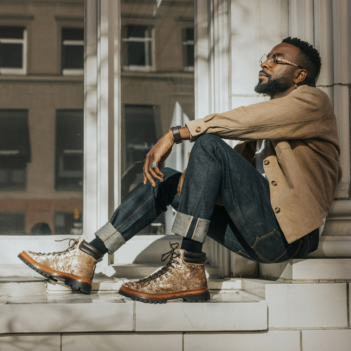 A man sits on a window ledge in casual attire. He wears glasses, a beige jacket, rolled-up jeans, and brown hiking boots.