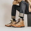 A person wearing denim jeans and beige suede hiking boots with black laces, sitting on a stool against a plain background.