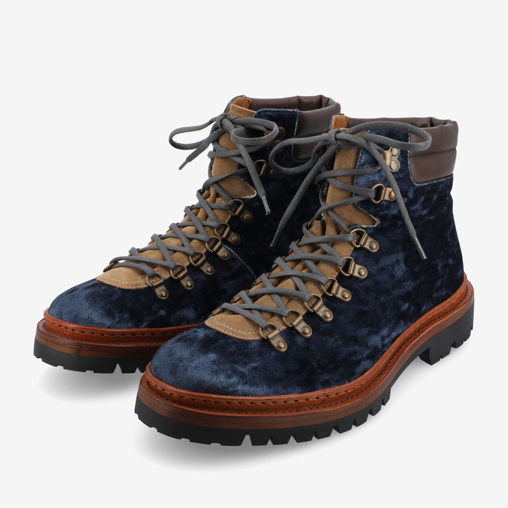 A pair of dark blue velvet hiking boots with brown leather trim, beige eyelet area, grey laces, and black textured soles on a white background.