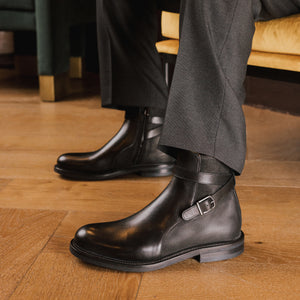 Dark leather boots with buckles worn by a person sitting on a wooden chair.