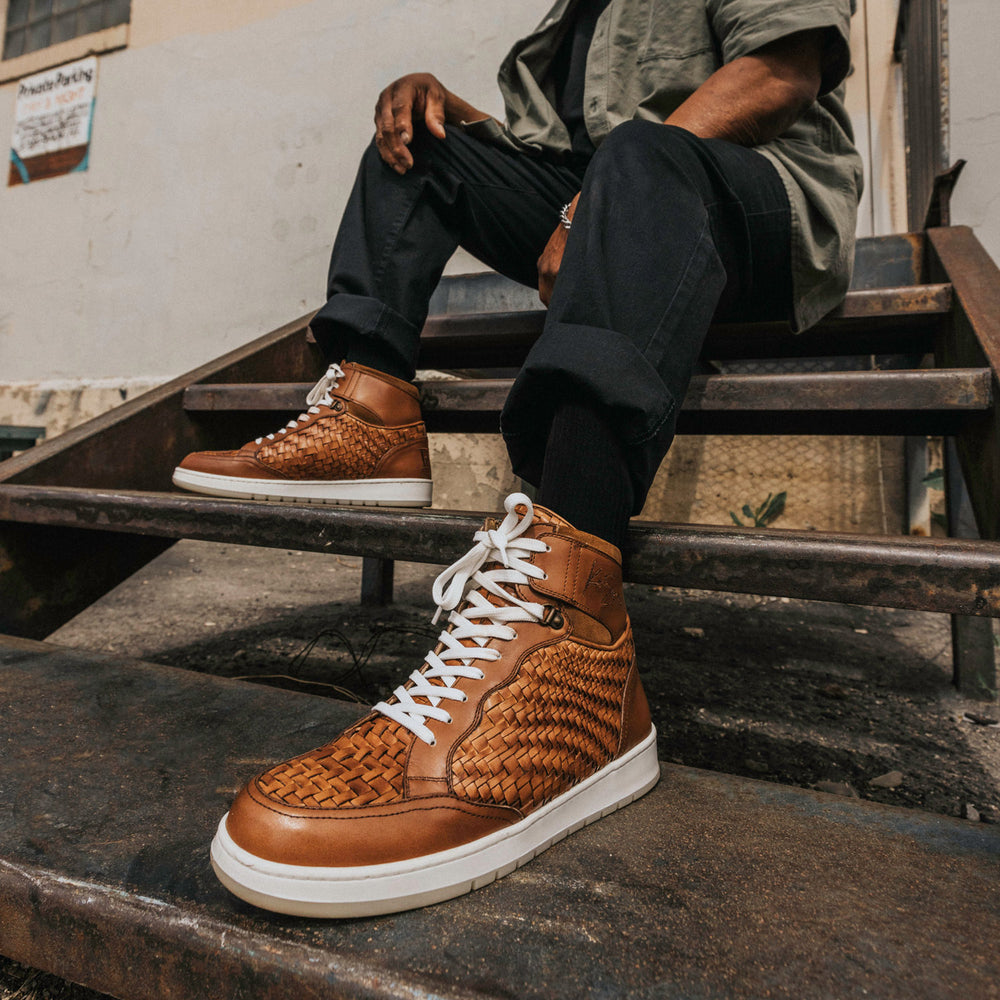 A person is sitting on outdoor metal stairs, wearing black pants, a green shirt, and brown woven high-top sneakers with white soles and laces.