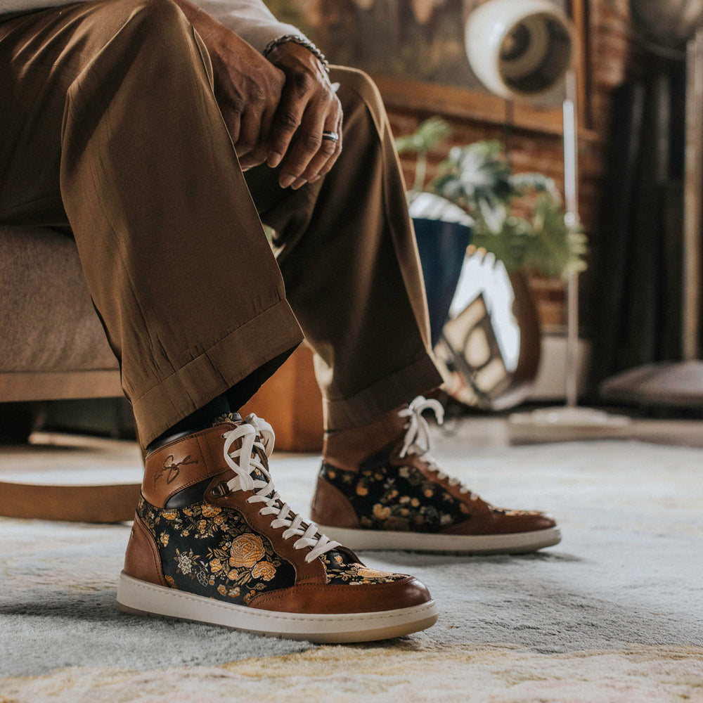 Person wearing brown pants and patterned sneakers with white laces, sitting on a chair in a room with a rug and various decor items.