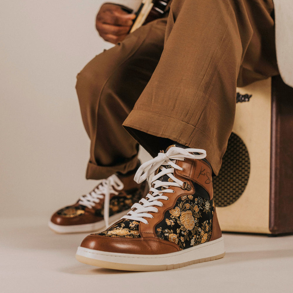 A person wearing brown trousers and floral high-top sneakers with white laces sits on or near an amplifier, holding a musical instrument.