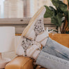 Person wearing white designer sneakers with a floral pattern, relaxed on a tan couch. The jeans are light blue and cuffed.