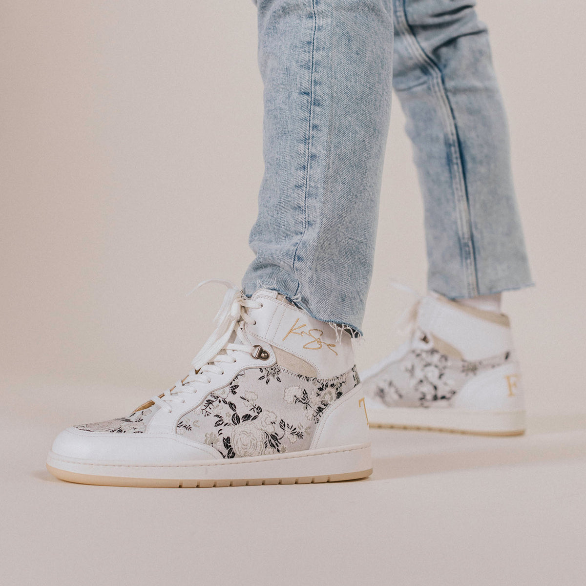 Close-up of a person wearing light blue jeans and white high-top sneakers with a black floral pattern and golden accents.