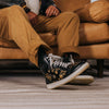 A person wearing tan pants is seated on a brown leather couch, showcasing black and gold floral patterned sneakers with white laces.