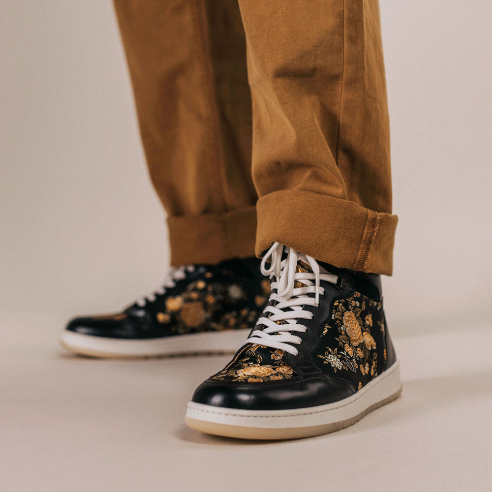 A person wearing brown pants and black high-top sneakers with an intricate gold floral design stands on a neutral background.