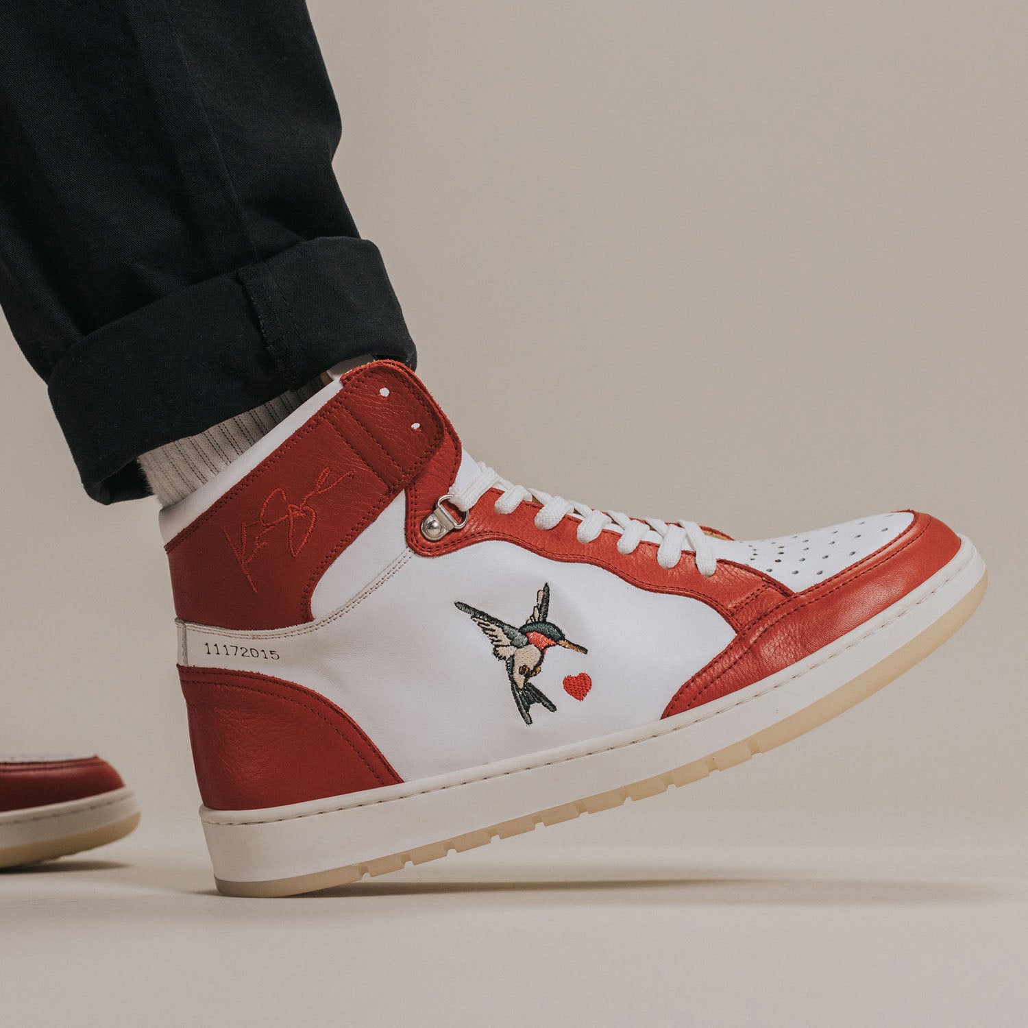 A close-up of a person wearing red and white high-top sneakers with a bird and heart design on the side, and rolled-up dark pants.