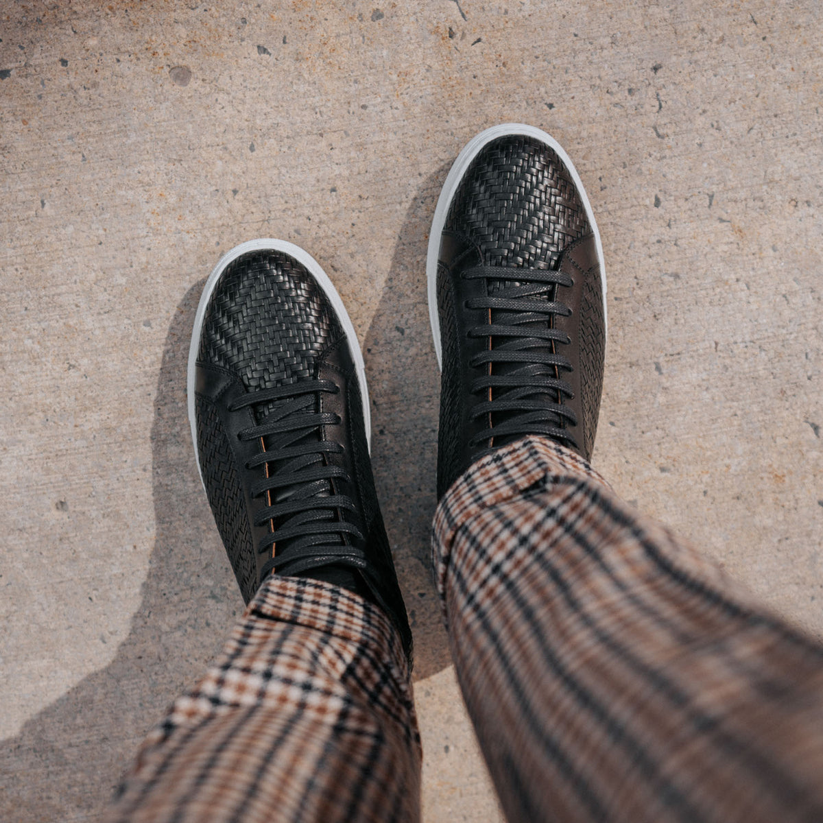 A person wearing black woven sneakers and brown plaid pants stands on a concrete surface.