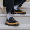 A person wearing black loafers with thick soles, white socks, and frayed-hem black pants sits on a metal staircase.