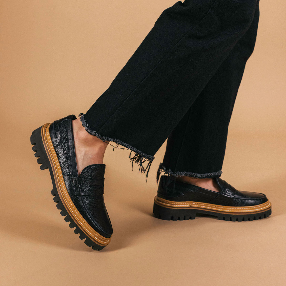 A person wearing distressed black jeans and black loafers with thick brown soles poses against a beige background.