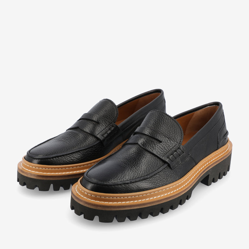 Black leather loafers with rugged, thick soles and tan midsoles. The shoes have a slip-on design and visible stitching details.