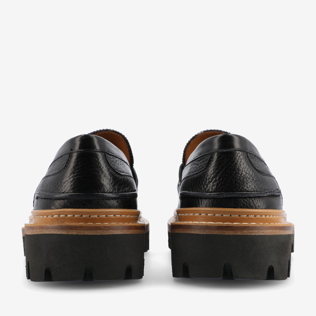 Rear view of black leather loafers featuring a pebbled texture, brown midsole, and thick black rubber outsoles with deep treads.