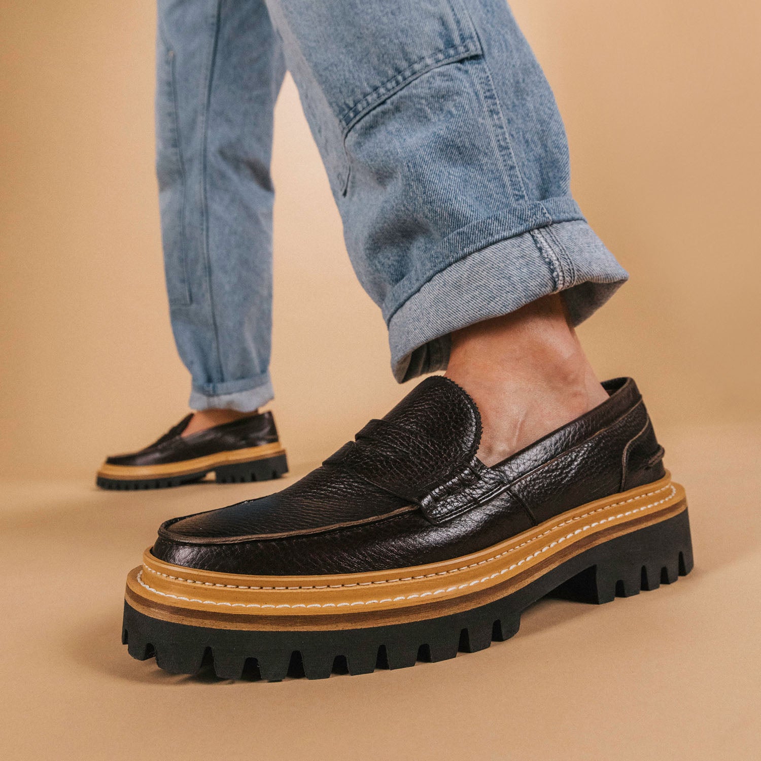 Person wearing brown leather loafers with thick, treaded soles and light brown accents, combined with light blue rolled-up jeans against a beige background.