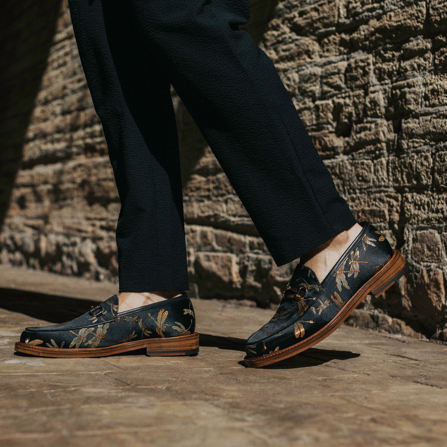 A person wearing black pants and navy blue loafers with golden floral patterns walking on a brick-paved surface.