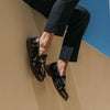 A person wearing black and gold embroidered loafers and black pants is seated on a beige wall against a clear sky.