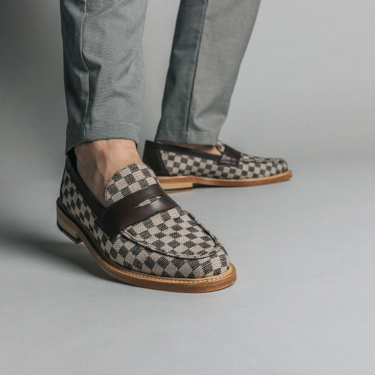 A person wearing gray pants and checkered loafers with dark leather straps standing on a light gray floor.