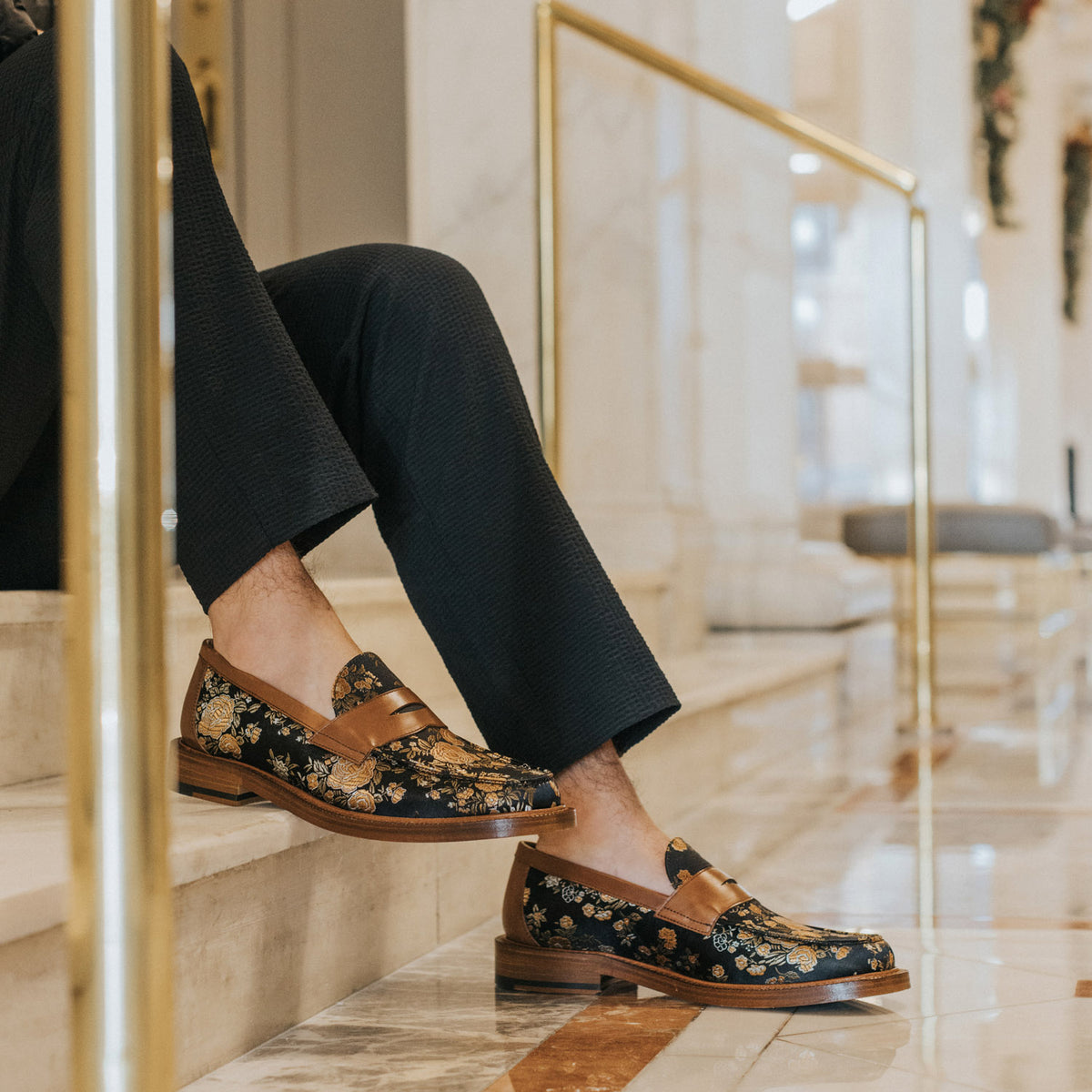 A person wearing black pants and floral-patterned loafers sits on a marble staircase, resting their feet on the step.