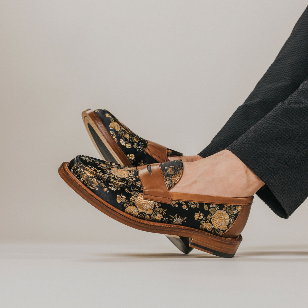 A person wearing black floral-patterned loafers with beige and gold accents, paired with dark trousers, stands on a light background.