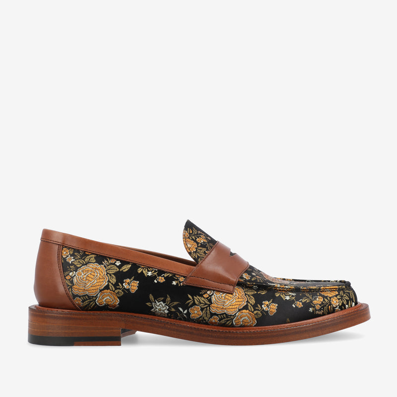 A black loafer with golden floral embroidery and brown leather accents on the heel and panels. The shoe has a wooden sole with a slight heel.