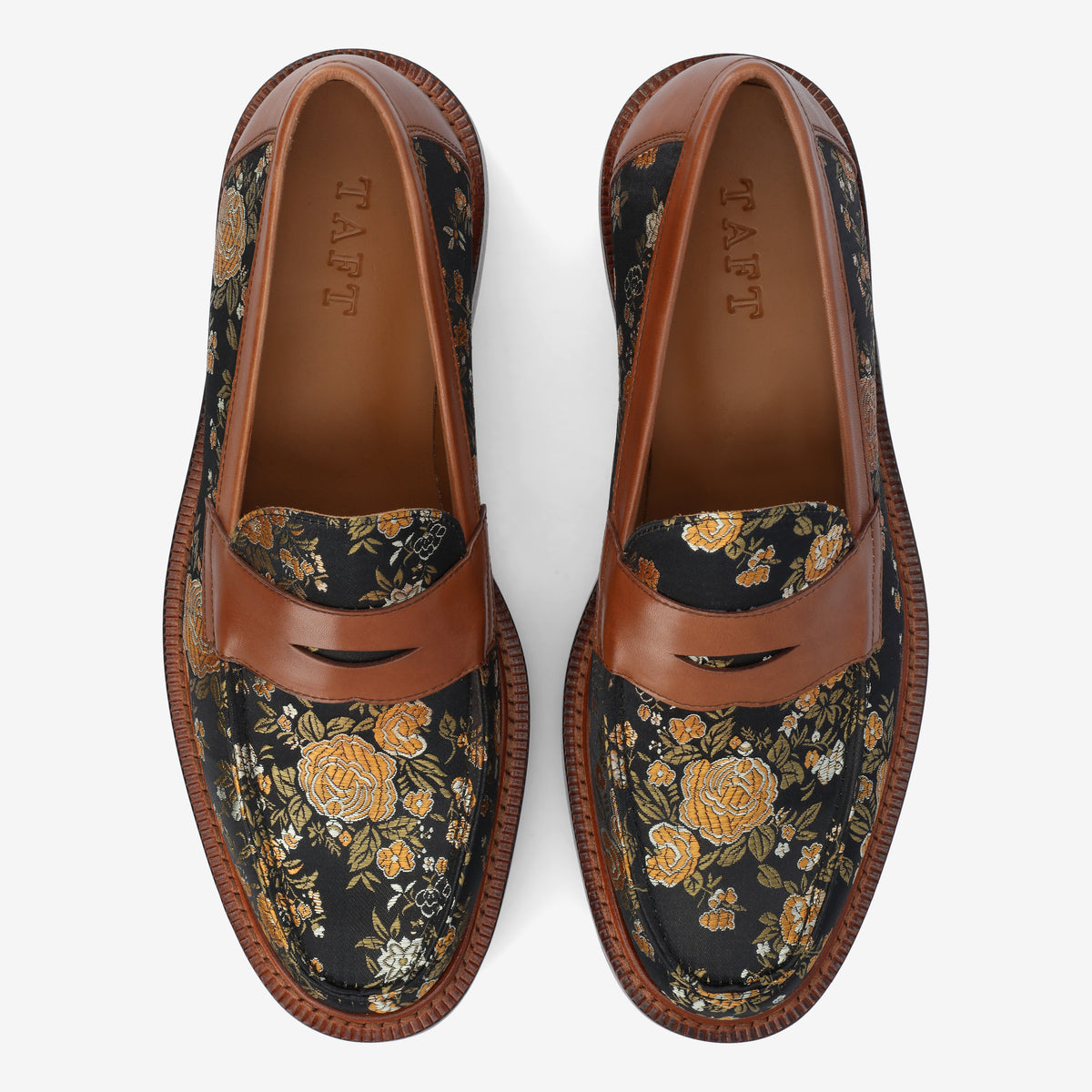 A pair of men's loafers featuring a black fabric with yellow and orange floral patterns, brown leather straps, and brown trim. The insole reads TAFT.