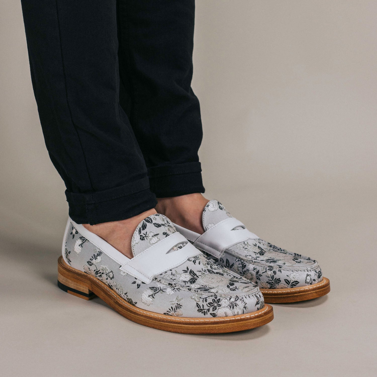 A person wearing black pants and white loafers with a black floral pattern stands on a neutral background.