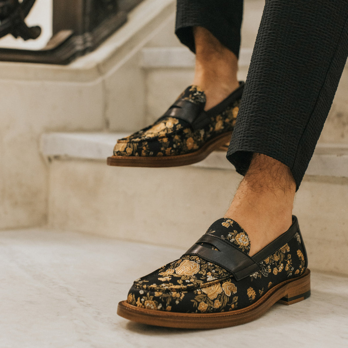 A person wearing black and gold floral-patterned loafers descends a marble staircase.