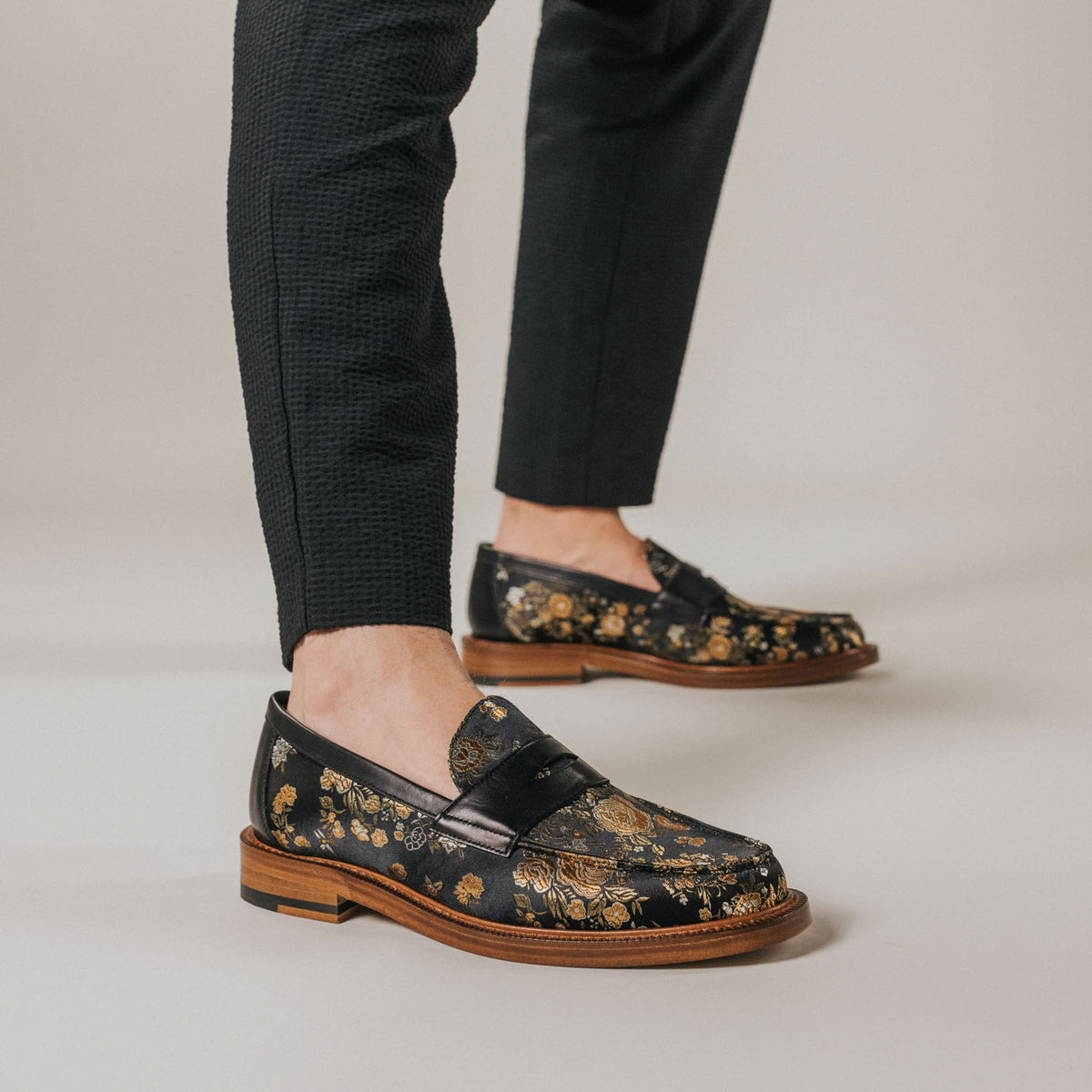 Person wearing black dress pants and black loafers with a gold floral pattern, standing on a neutral background.