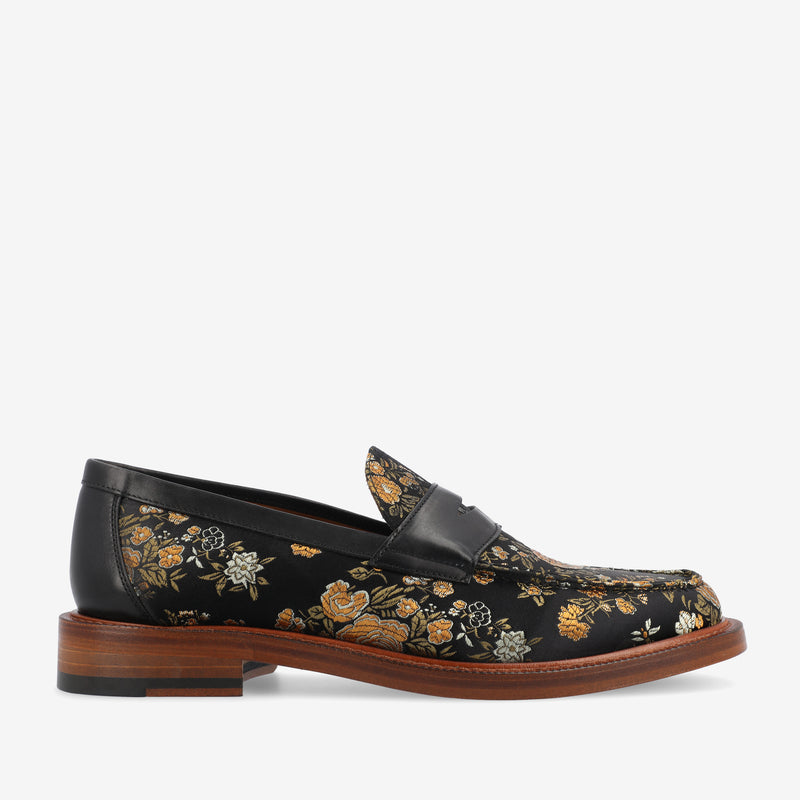 A black leather loafer with a floral pattern in gold and white, and a brown heel.