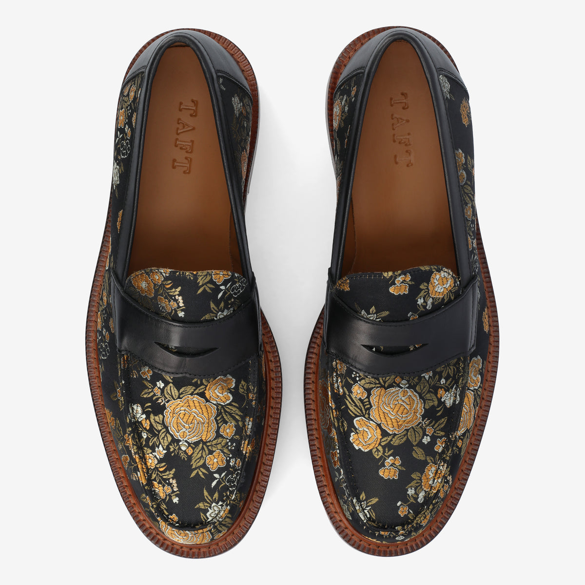 A pair of black loafers with a floral pattern in yellow and green, featuring brown stitched soles. The word TAFT is embossed inside the shoes.