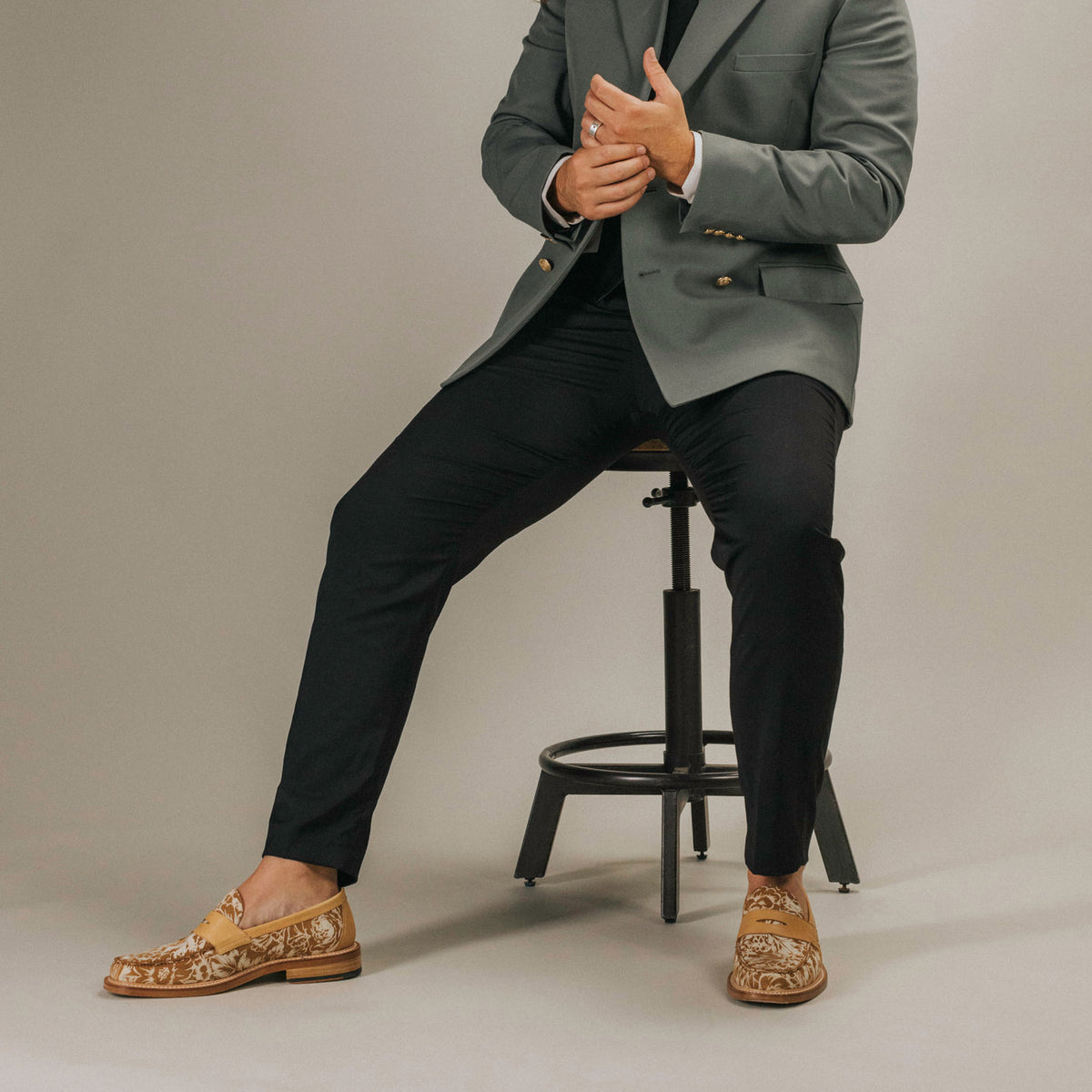 A person sits on a stool, dressed in a gray blazer, black pants, and brown patterned loafers.