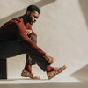 A man sits on a stool adjusting one of his floral-patterned shoes, wearing a red sweater, dark pants, and red socks, with a neutral background.