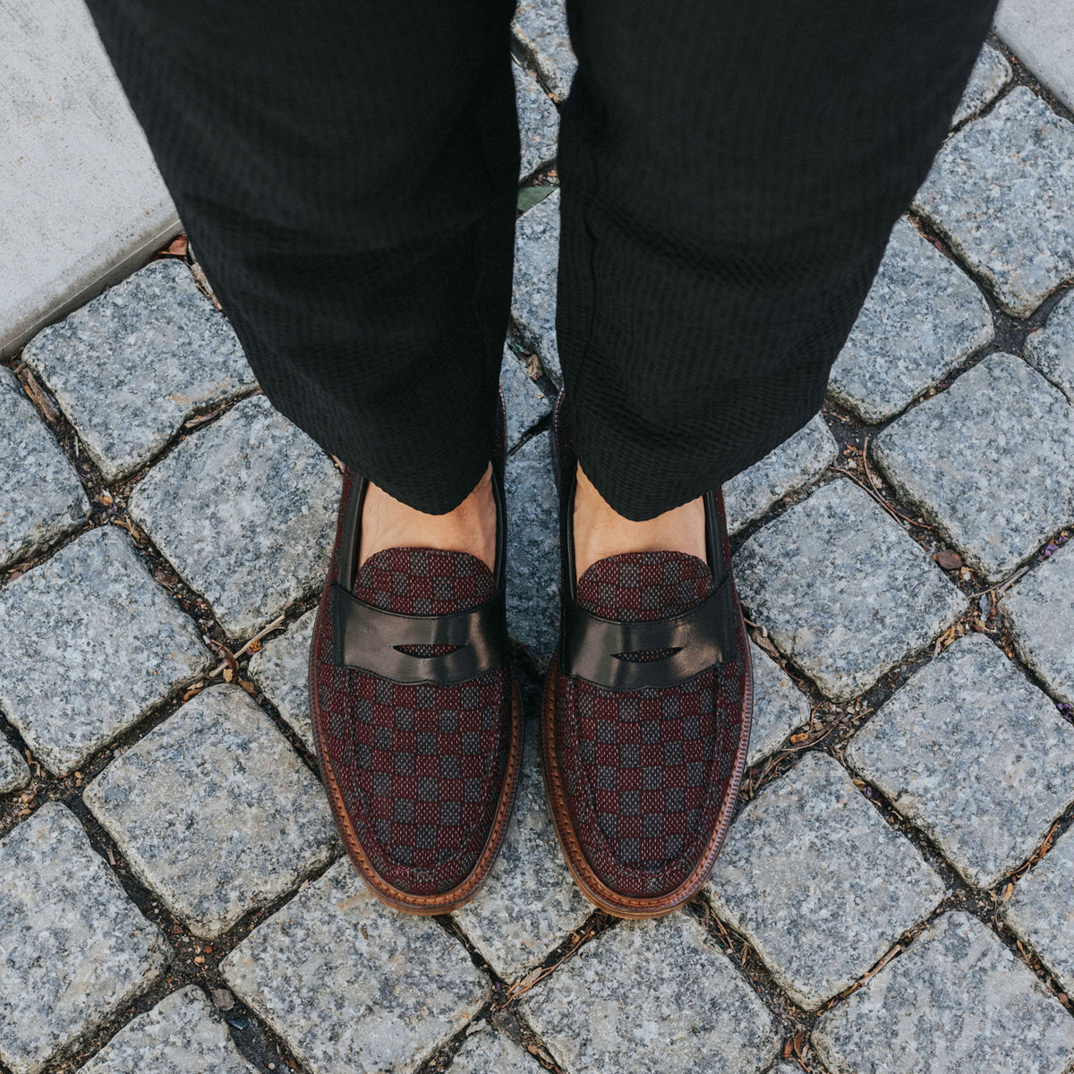 A person wearing black pants and dark red checkered loafers with black leather straps stands on a cobblestone pavement.